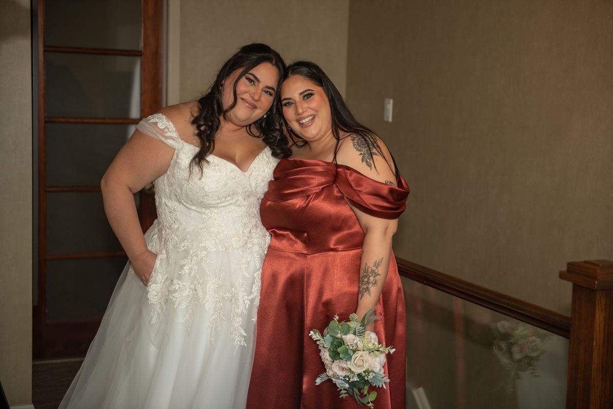 Two brides in joyful poses, one in white lace and the other in rust satin with pastel flowers, stand on a staircase.