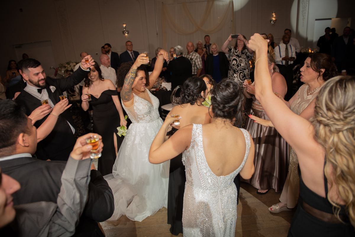 Guests including the bride in a white gown dance and raise glasses at a wedding reception under warm ambient lighting.