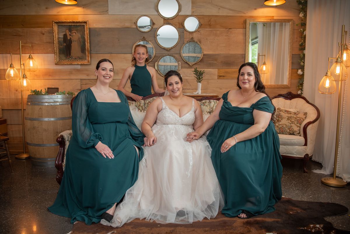 A bride and two bridesmaids in elegant dresses smile warmly in a rustic wooden room with golden lighting.