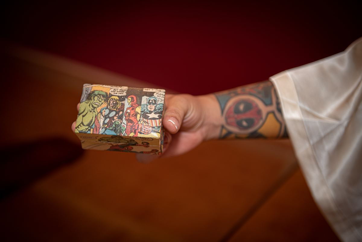 A person in a white robe offers a comic-book-patterned box with a tattooed hand against a warm red background.