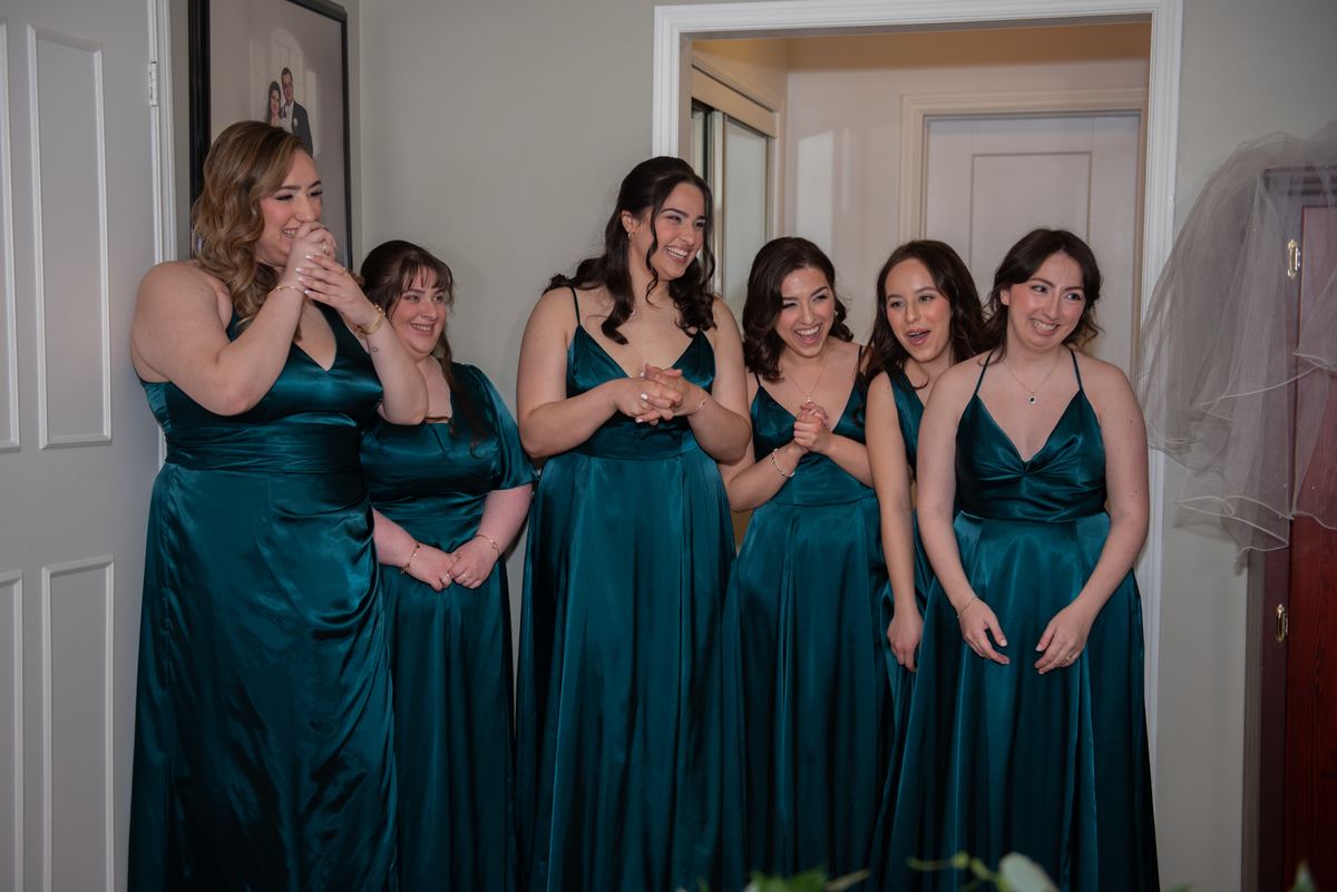 Bridesmaids in emerald satin gowns laugh joyfully in a hallway, illuminated by soft natural light from an interior doorway.