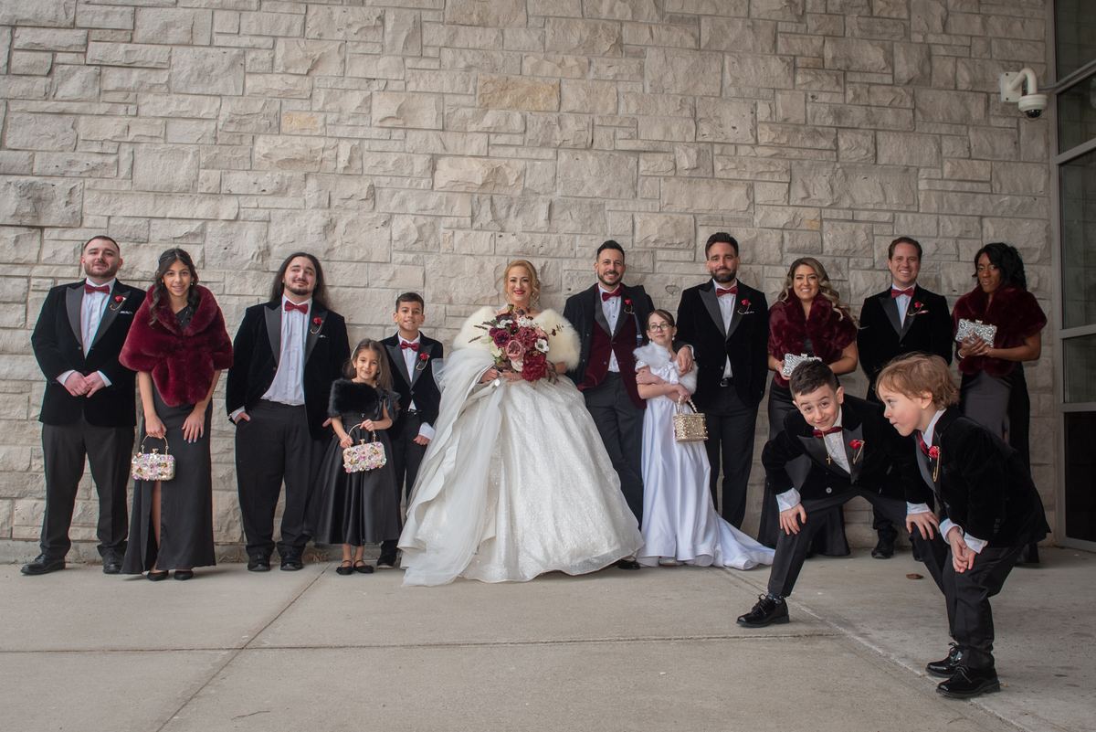 A formal wedding party poses outdoors against a stone wall, dressed in tuxedos and gowns with burgundy accents.