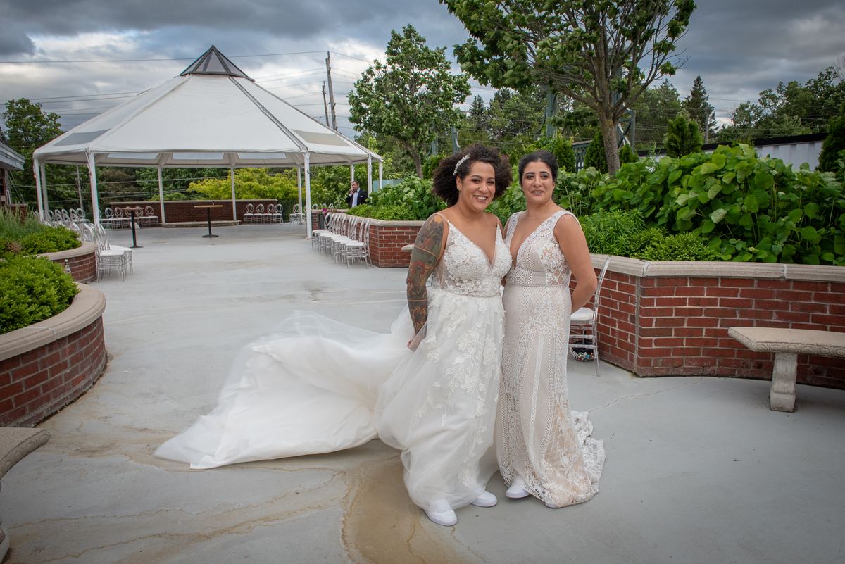 Two brides in lace wedding gowns smile at the camera in a courtyard surrounded by lush greenery and a white tented gazebo.