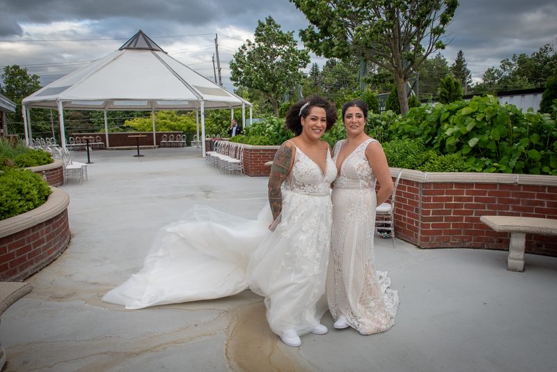 Two brides in lace wedding gowns smile at the camera in a courtyard surrounded by lush greenery and a white tented gazebo.