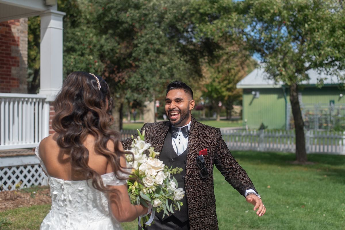 Groom in patterned tux laughing as bride approaches with white flowers on grassy lawn with trees and fence.