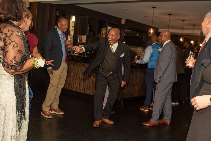 A bald man in a dark suit laughs and gestures toward someone off-camera at a festive wedding reception.