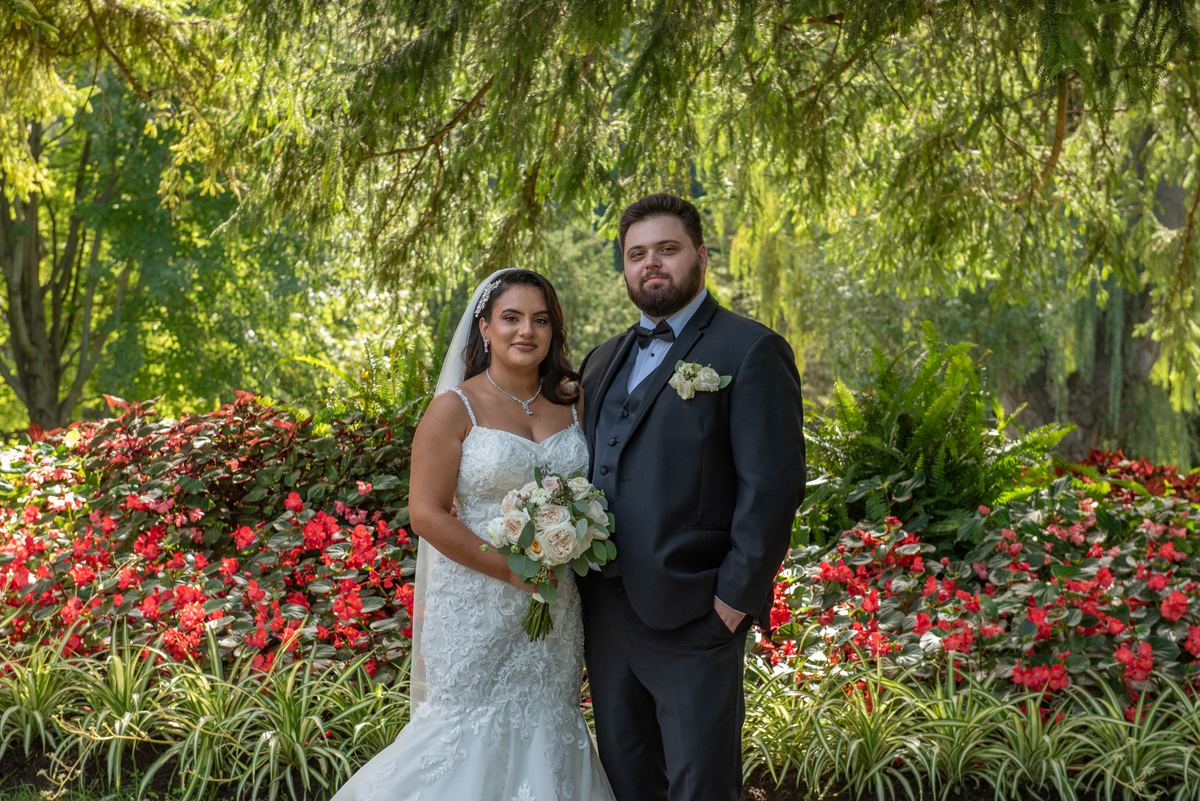 A bride in white and a groom in black stand smiling in a garden filled with red flowers under dappled sunlight.