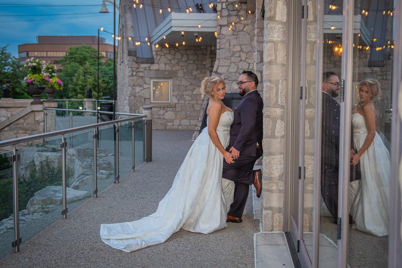 Bride and groom smile beside stone pillar, reflected in glass door as dusk light and fairy lights create romantic ambiance.