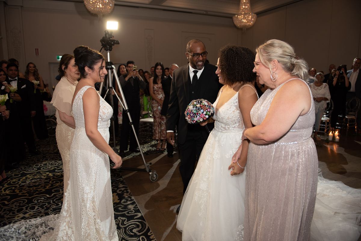 Two brides in elegant gowns walk toward each other in a grand hall with chandeliers and guests watching.