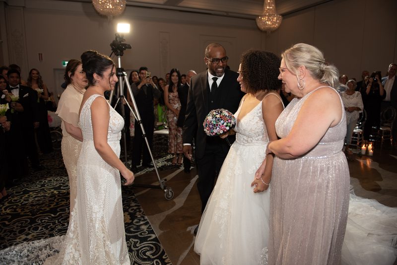 Two brides in elegant gowns walk toward each other in a grand hall with chandeliers and guests watching.