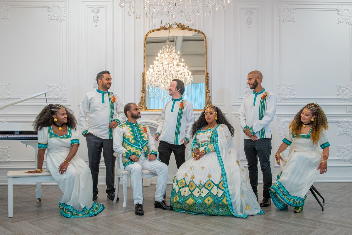 Seven people in traditional African attire pose joyfully in an elegant room with a grand chandelier.