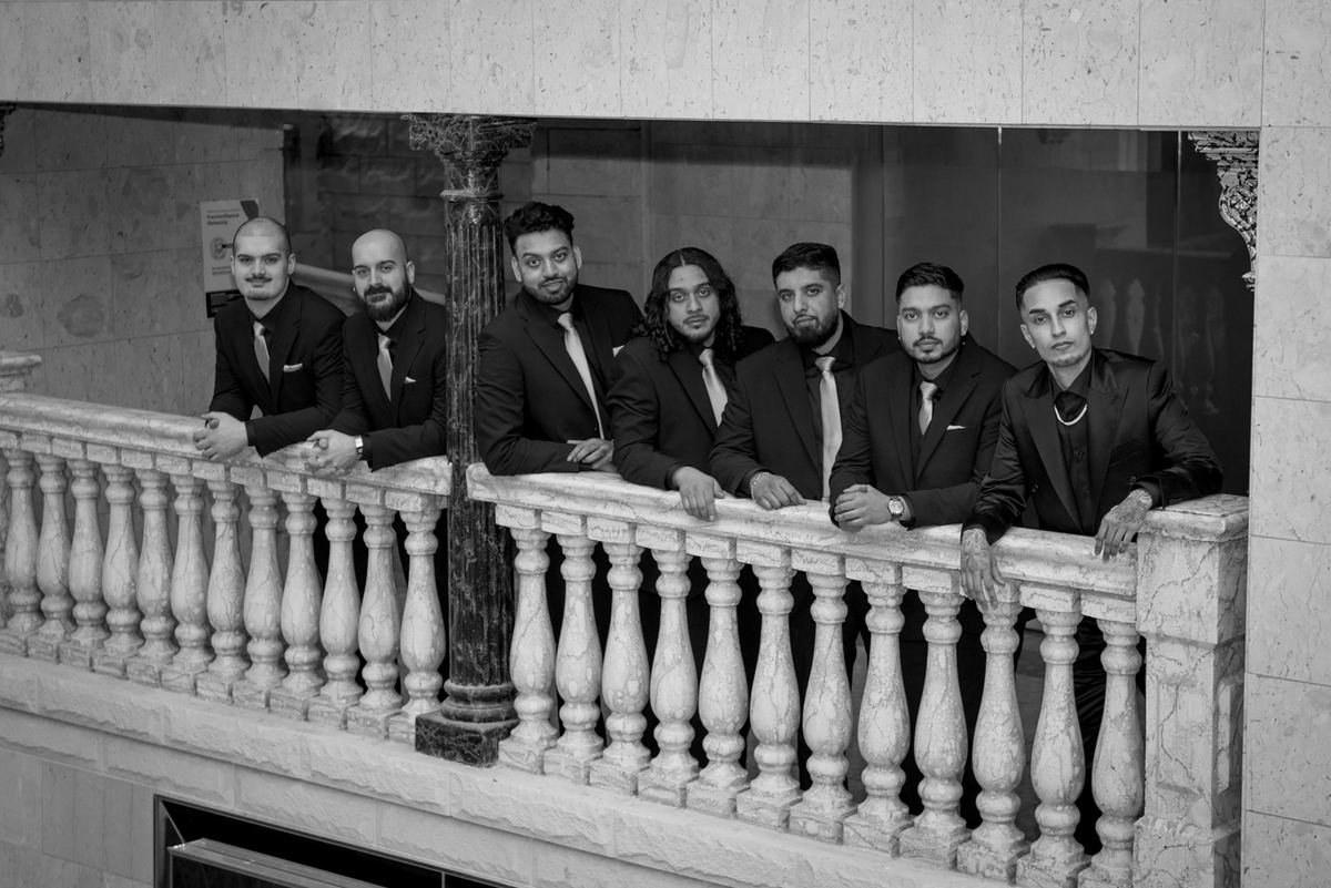 Seven formally dressed men stand together on a marble balcony, leaning on the balustrade and looking at the camera.