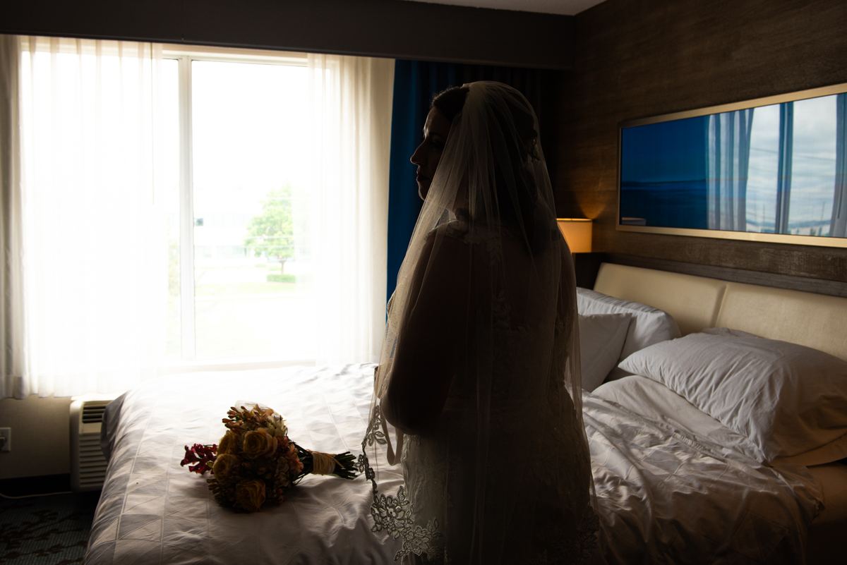 A bride stands beside a hotel bed where her bouquet rests, silhouetted against the bright window light.