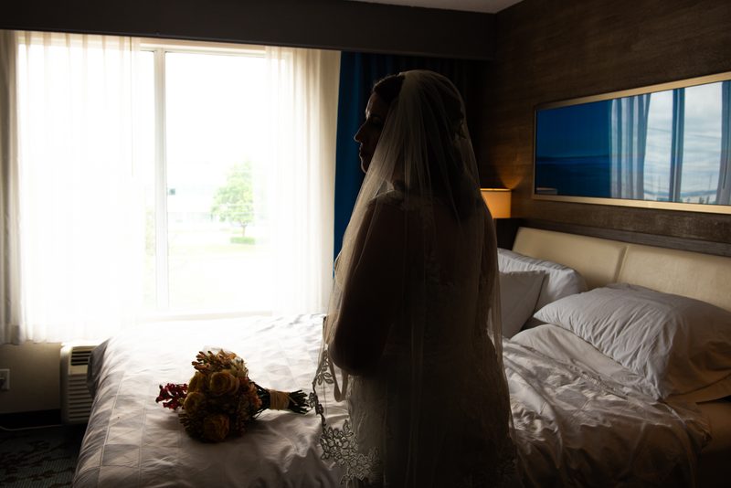A bride stands beside a hotel bed where her bouquet rests, silhouetted against the bright window light.