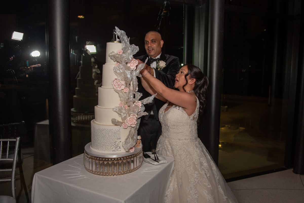 Bride and groom cut their multi-tiered wedding cake with pink floral accents, smiles reflecting the bright lights.