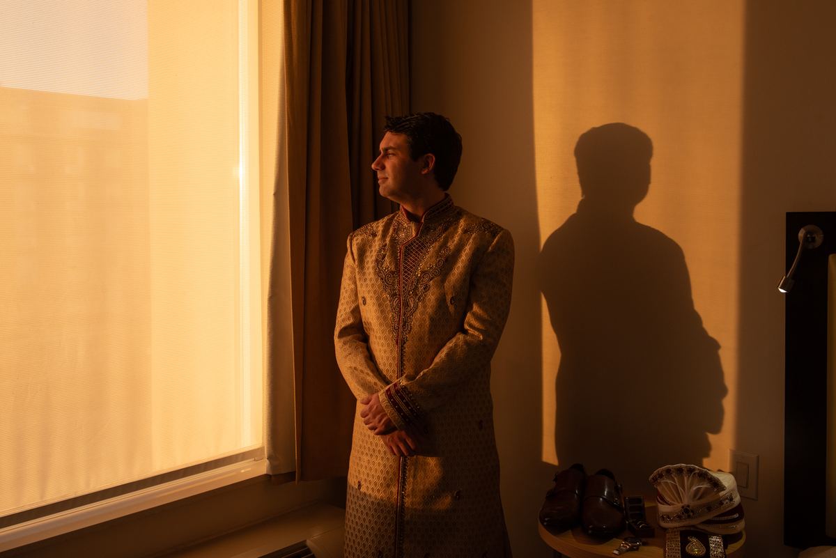 A man in a traditional Indian sherwani stands by a window, light casting a long shadow on the wall beside him.