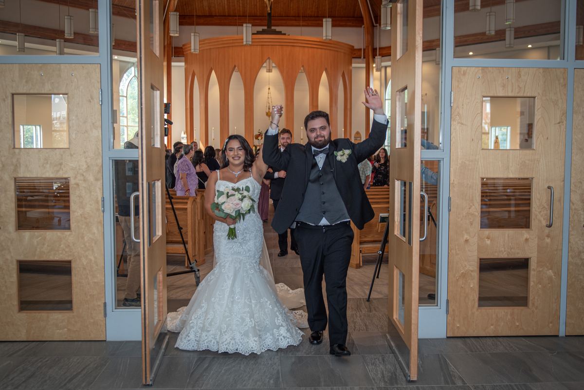 A bride and groom exit a modern church, waving to guests as natural light fills the warmly lit interior with wooden beams.