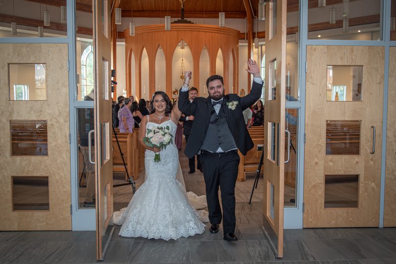A bride and groom exit a modern church, waving to guests as natural light fills the warmly lit interior with wooden beams.