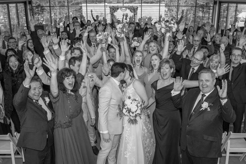 Amanda and Justin stand at the center of a joyous celebration surrounded by guests raising their hands in cheer.