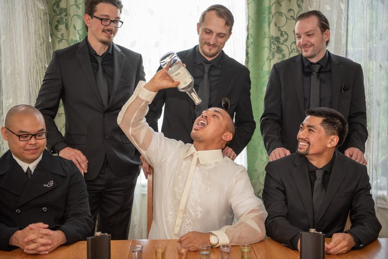 A groom pours liquor into his mouth while surrounded by smiling groomsmen in black suits, celebrating.