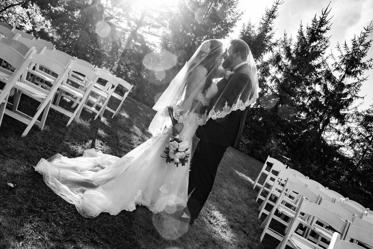 A bride and groom kiss outdoors, surrounded by white chairs and trees under a bright sky with lens flares.