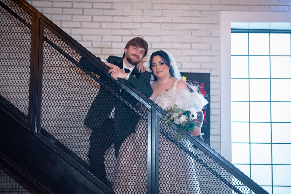 A bride and groom lean against a black metal railing on a staircase, illuminated by soft light from a large window.