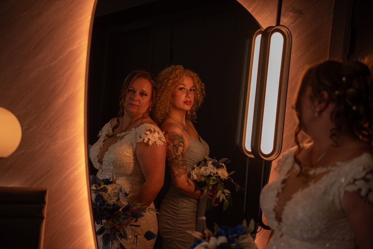 A bride in a white gown and a bridesmaid in a gray dress stand before a mirror, gazing at the camera in a warmly lit room.