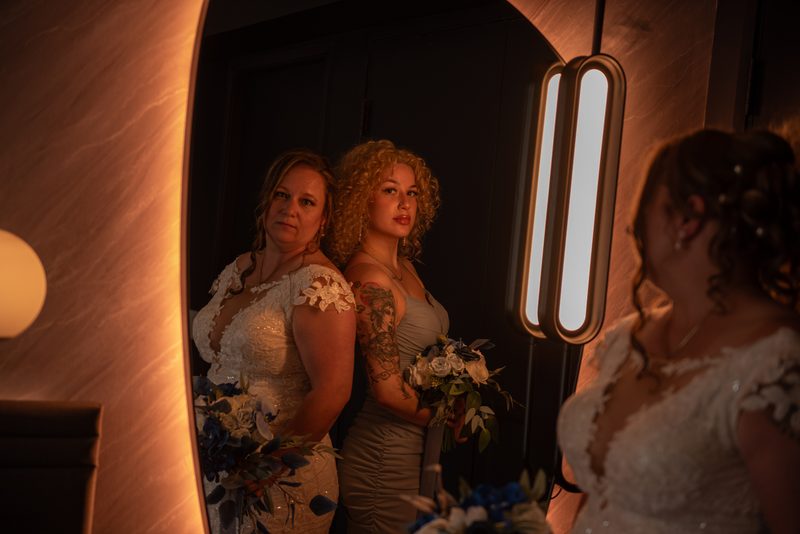 A bride in a white gown and a bridesmaid in a gray dress stand before a mirror, gazing at the camera in a warmly lit room.