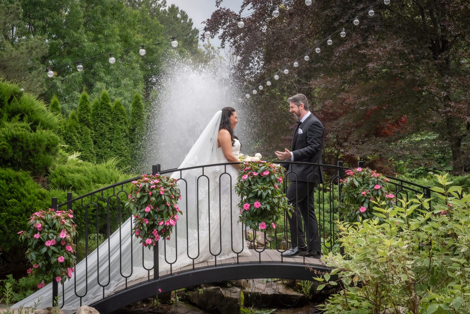 Bride and groom stand on a garden bridge, water spraying between them as pink flowers adorn the railing.