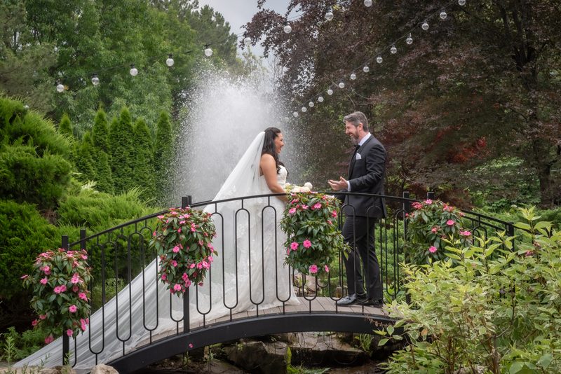 Bride and groom stand on a garden bridge, water spraying between them as pink flowers adorn the railing.