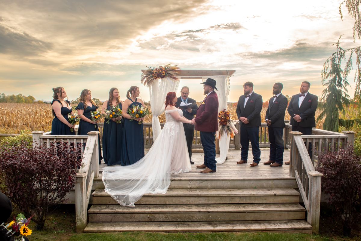 Bride and groom exchange vows at sunset under an autumnal flower-adorned arch with their wedding party.