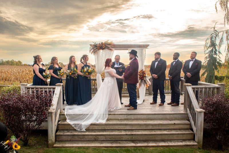 Bride and groom exchange vows at sunset under an autumnal flower-adorned arch with their wedding party.