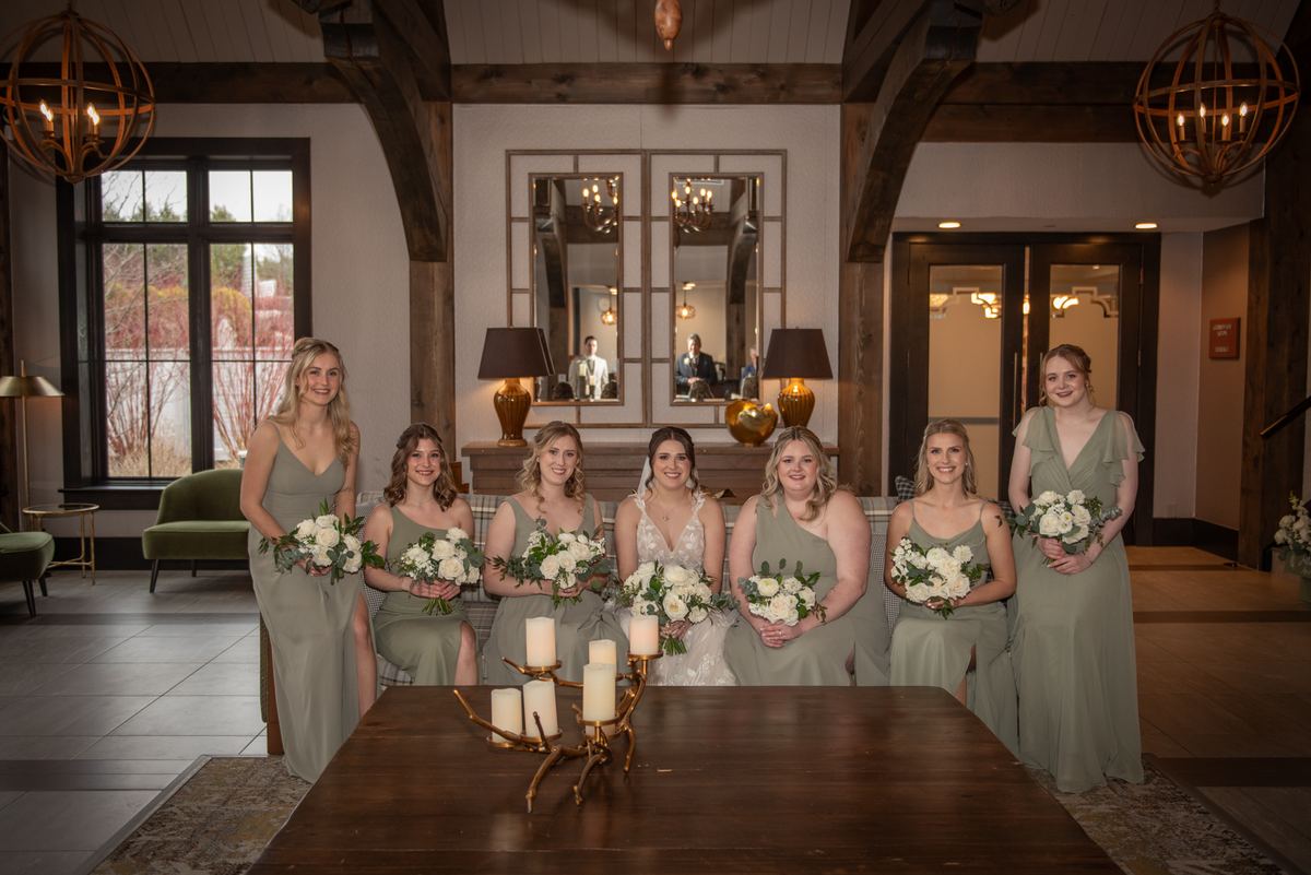 Bride and bridesmaids in sage green gowns with white flowers pose in rustic indoor space with exposed beams and chandeliers.
