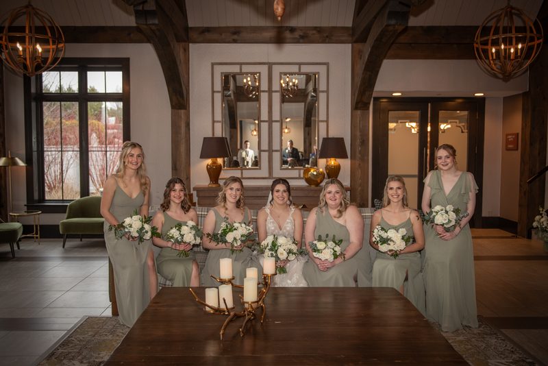 Bride and bridesmaids in sage green gowns with white flowers pose in rustic indoor space with exposed beams and chandeliers.