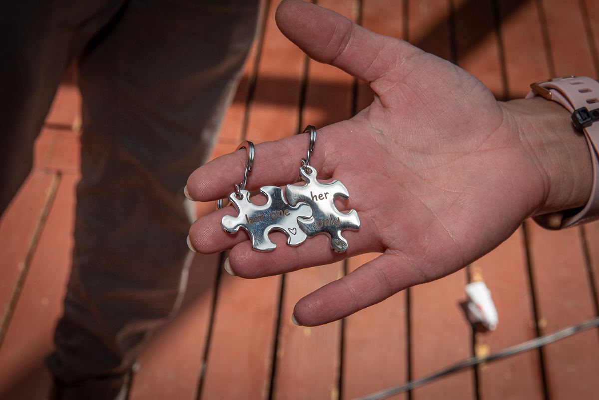 A hand holds interlocking puzzle-piece keychains engraved with "her" and "his," illuminated by sunlight on a wooden deck.
