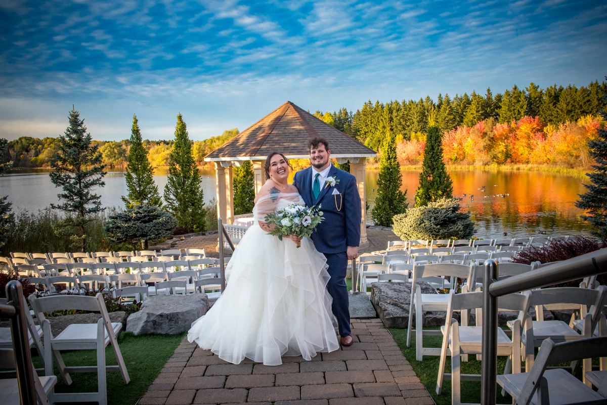 A smiling bride and groom stand on a brick path by a lake, surrounded by autumn trees and white chairs.