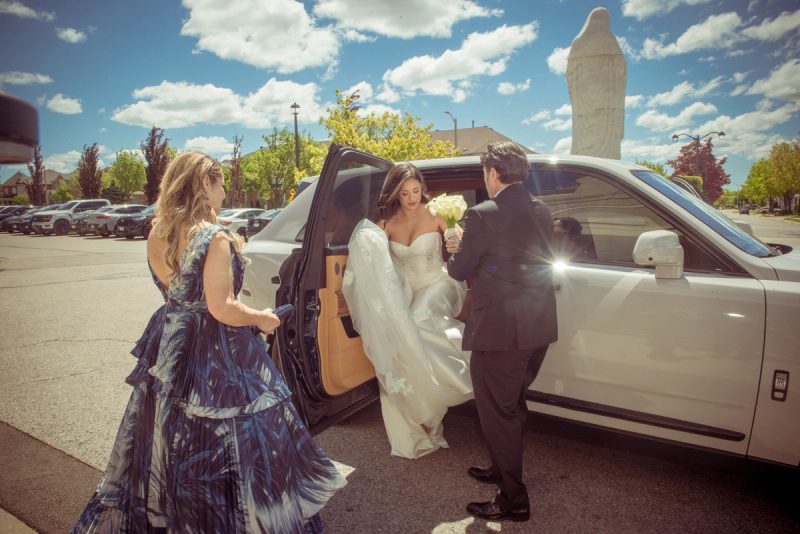 A bride exits a car with assistance as sunlight creates lens flares and highlights a statue in the background.