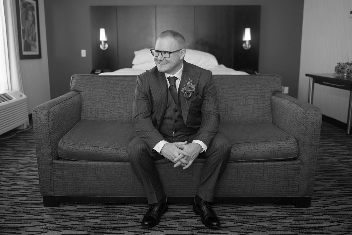 A groom in a pinstripe suit sits on a hotel sofa, smiling at the camera as natural light highlights his face.