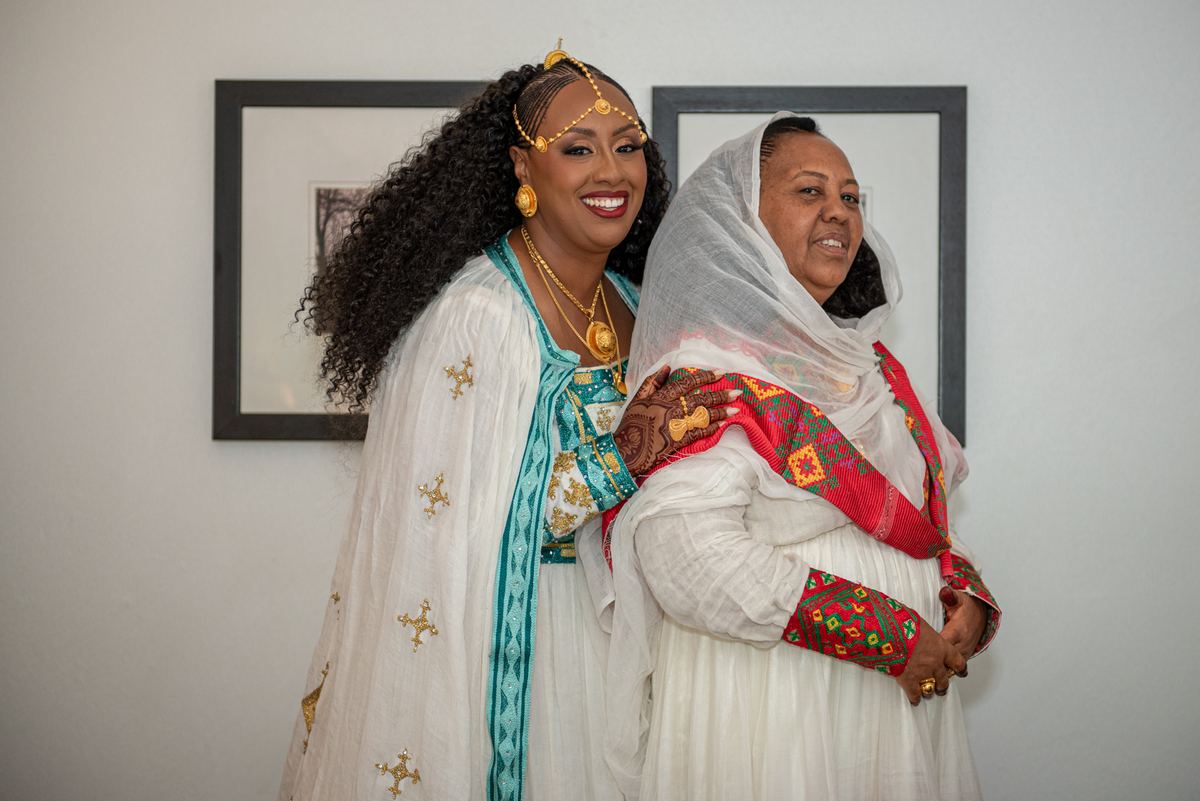 Two women in traditional attire stand close, one smiling brightly, the other offering support with a gentle touch.