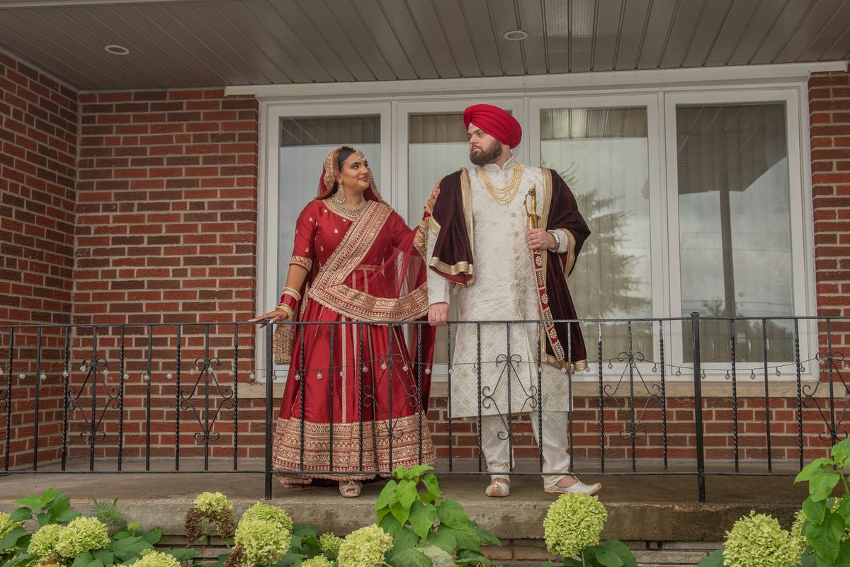 A newlywed couple in traditional South Asian attire stands on a porch, surrounded by green hydrangeas and a brick building.