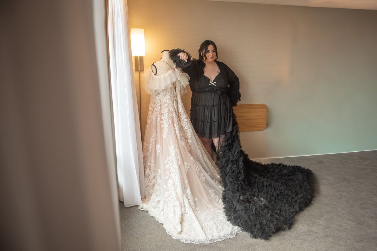 A woman stands beside a mannequin displaying an elaborate lace wedding gown, gently touching the fabric.