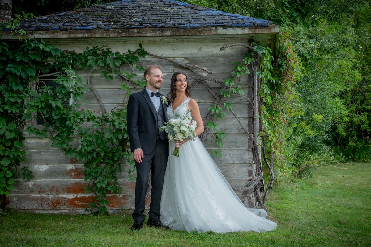 A smiling bride and groom stand beside an ivy-covered shed in a garden, illuminated by natural light.