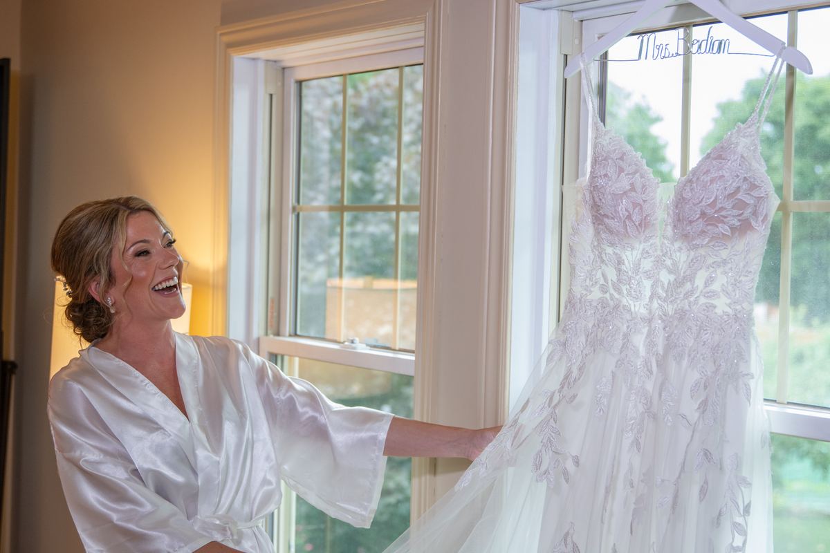 A bride in a satin robe laughs at her wedding gown hanging by the window, touching its lace detailing.