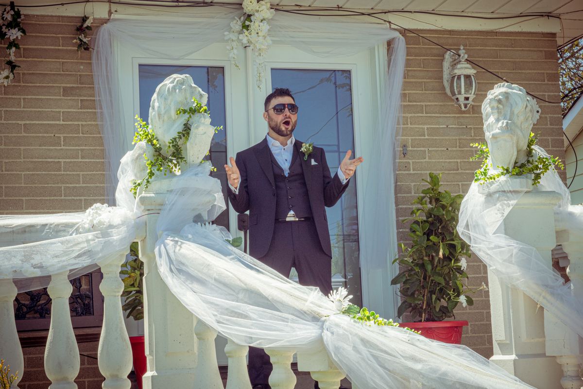 A man in a dark suit and sunglasses gestures enthusiastically on a decorated outdoor staircase during a celebration.