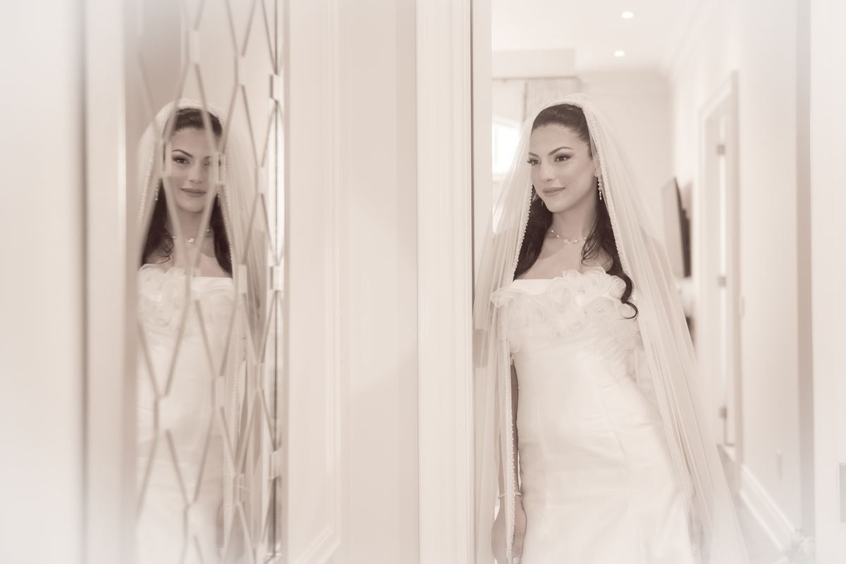 A bride in her wedding gown and veil stands thoughtfully in front of a mirrored door with geometric patterns.