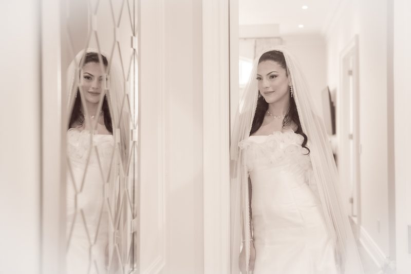 A bride in her wedding gown and veil stands thoughtfully in front of a mirrored door with geometric patterns.