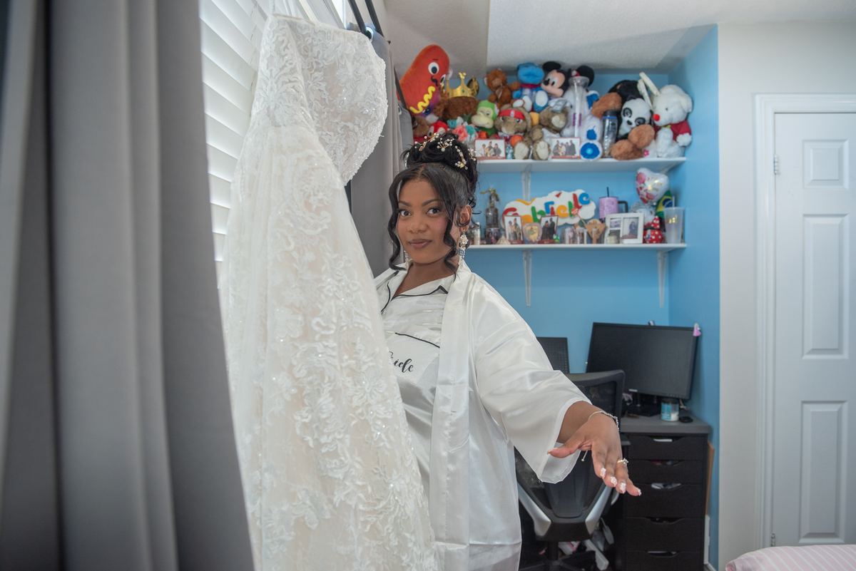 A bride gestures toward her wedding gown hanging in her bedroom, filled with personal touches and soft light.