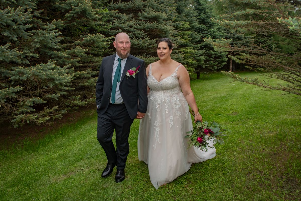 A smiling bride in a lace-trimmed gown and groom in a dark suit stand hand-in-hand on a grassy lawn surrounded by evergreens.