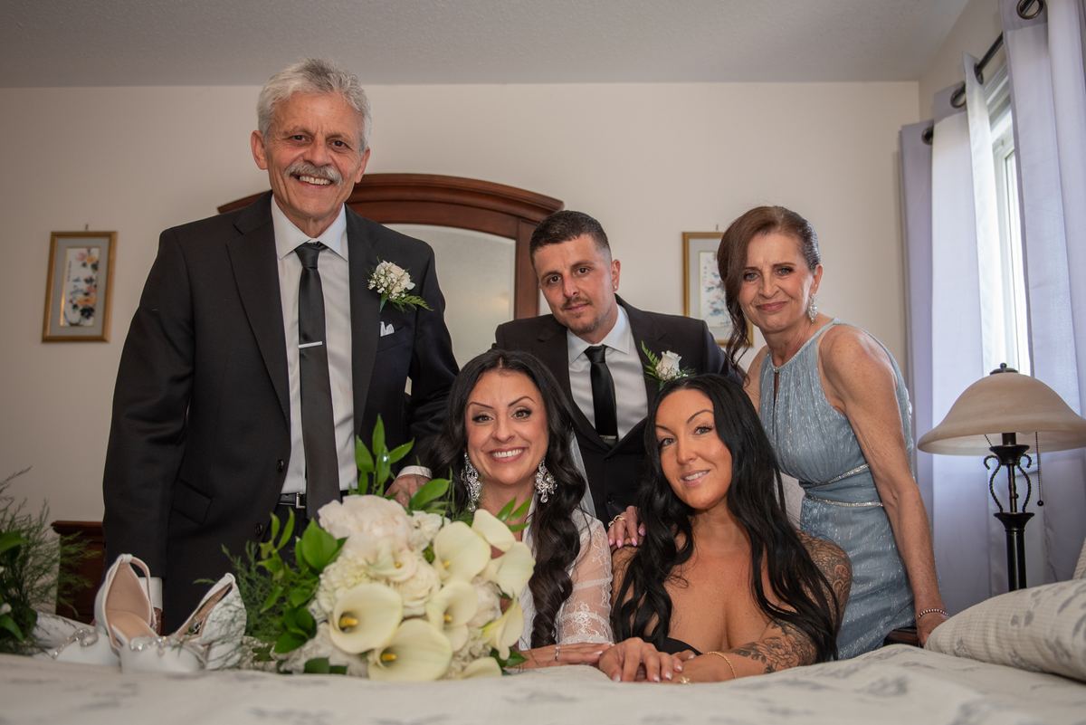 A bride and groom sit on a bed with their smiling parents in soft natural light during a wedding celebration.