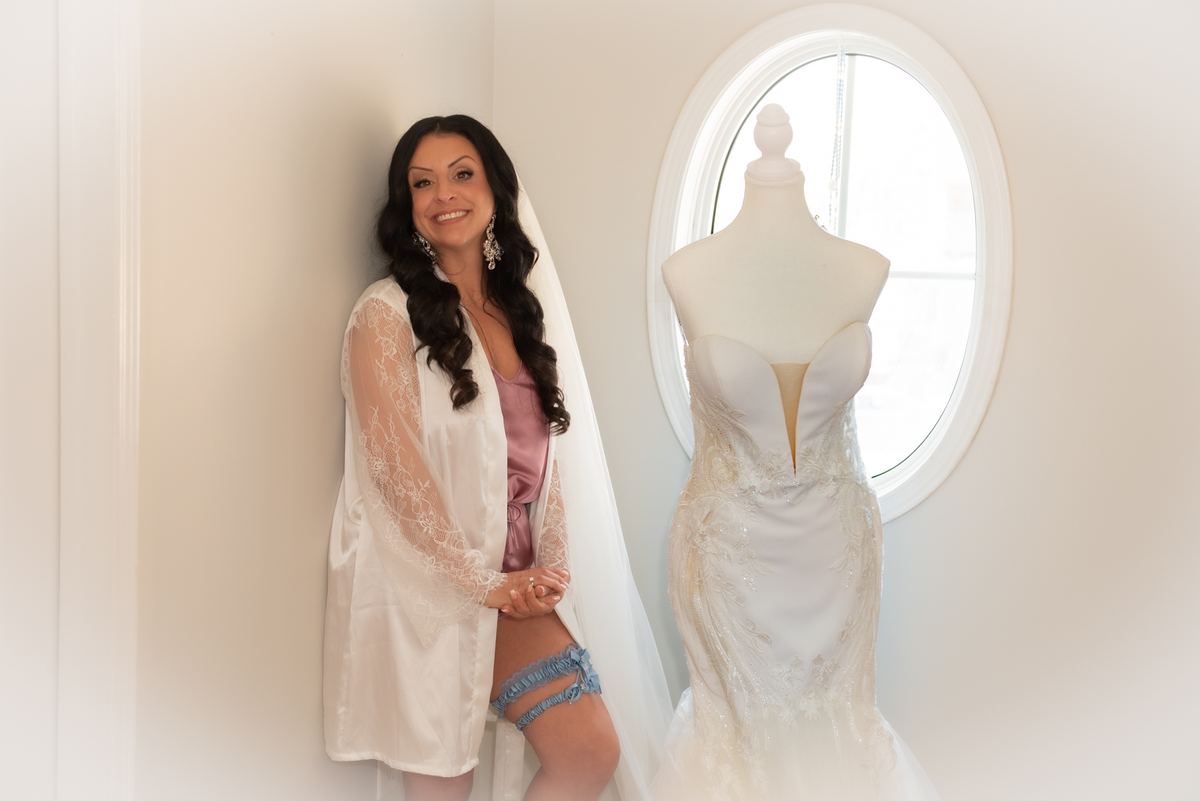 A bride in a lace robe and pink slip leans against a mannequin displaying her wedding gown beside an oval window.